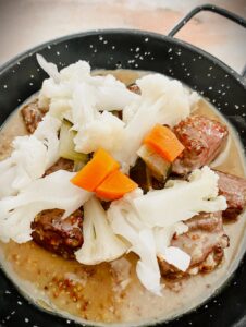 A black enamel bowl filled with a stew with pieces of meat then topped with pieces of cauliflower and carrots